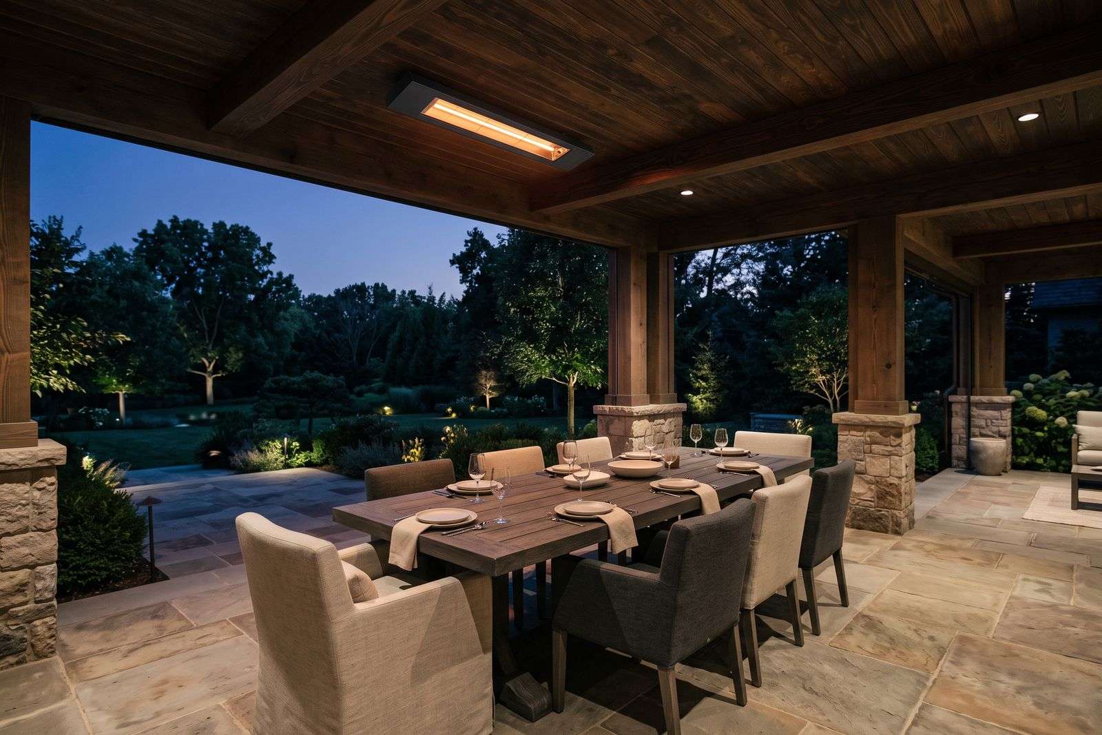 A slim matte-black heater mounted under the wood ceiling of a covered patio at dusk, glowing above an outdoor dining table.