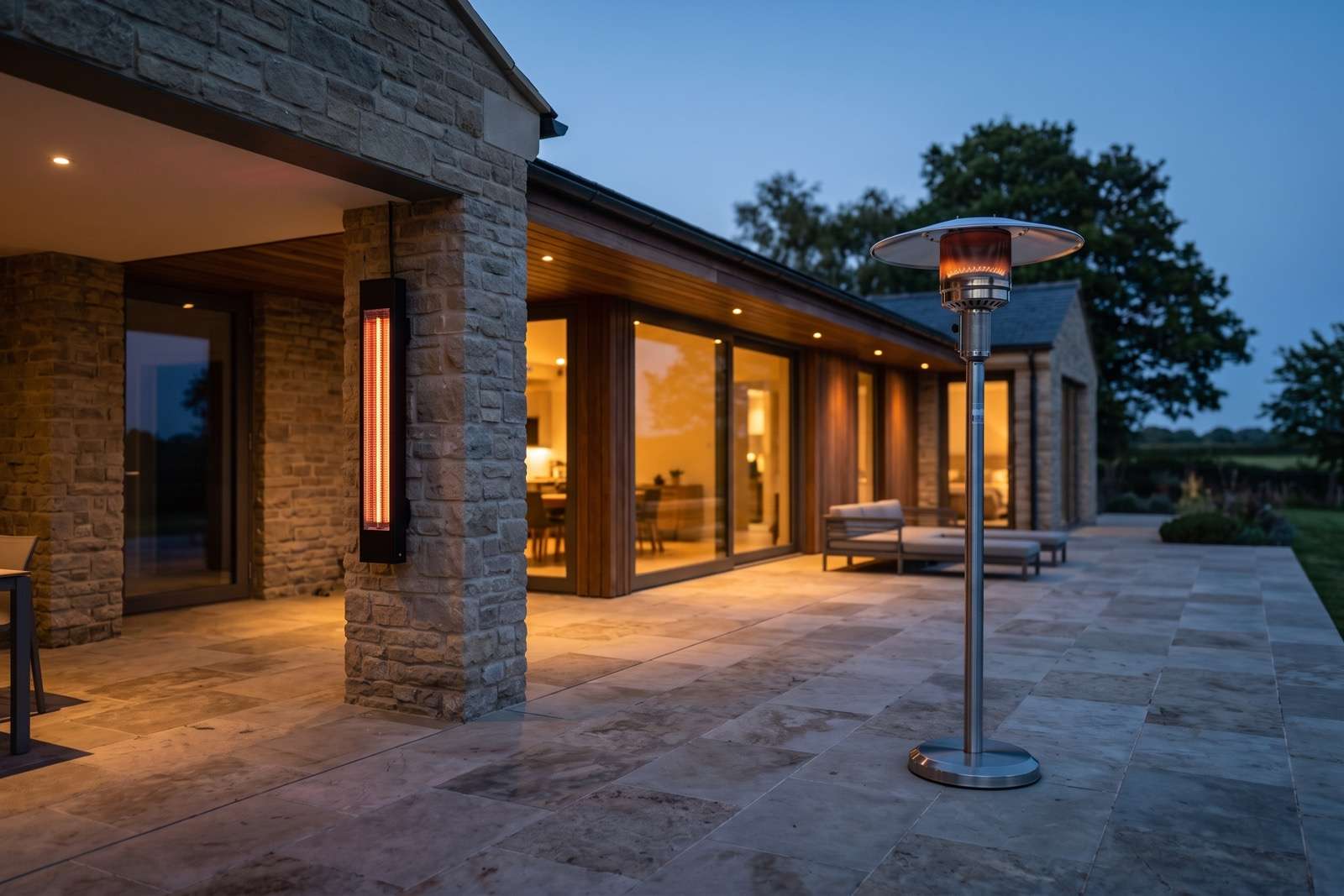 A wall-mounted electric infrared heater glowing under a patio roof beside a freestanding propane patio heater on an open stone terrace at dusk.
