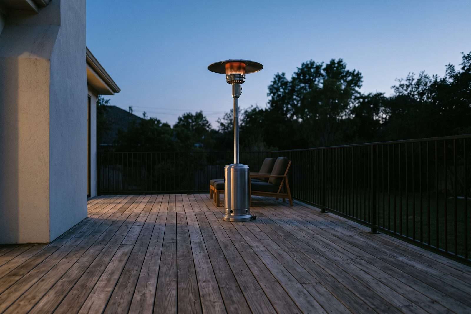 A wood deck at dusk with a tall stainless propane patio heater glowing near the middle of the deck, well clear of the house wall and the railing.