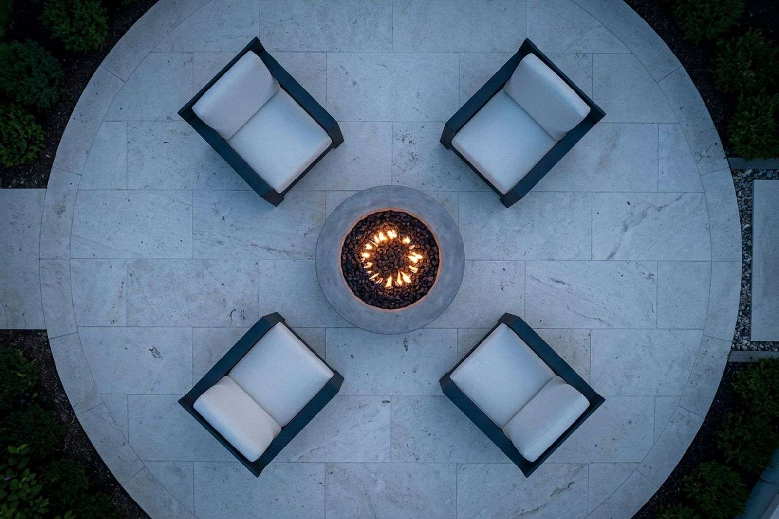 Overhead view of a round fire pit ringed by four lounge chairs on a stone patio at dusk.