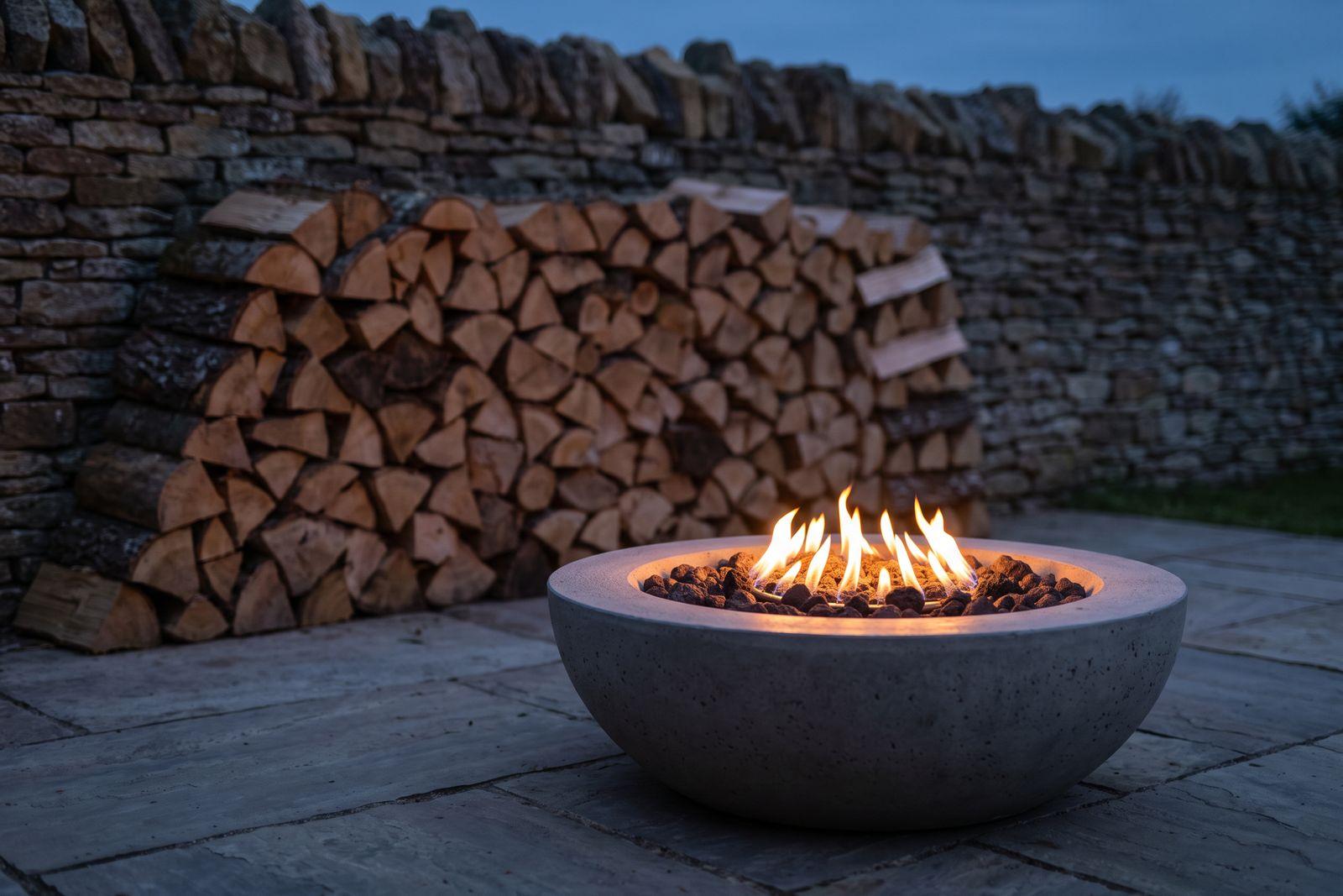 A gas fire bowl burning in the foreground with a stack of split firewood in soft focus behind it, on a patio at dusk.