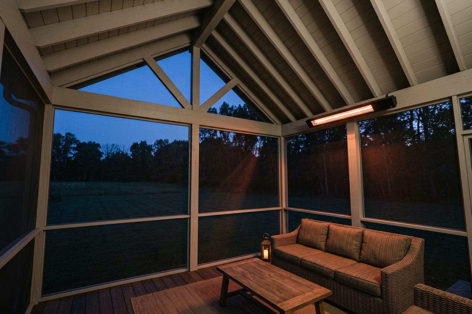 A screened porch at dusk with a slim black electric infrared heater mounted under the roof beam above a wicker sofa, its element glowing orange.