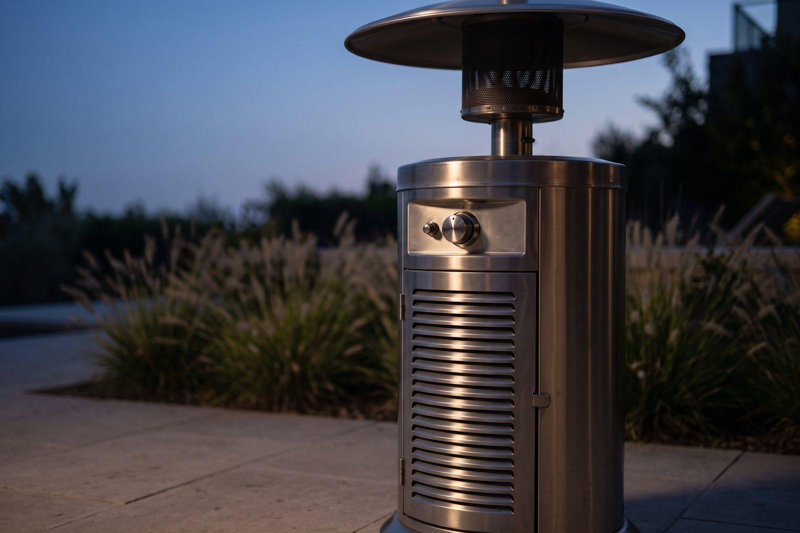 A freestanding propane patio heater at dusk on a stone patio, its control knob and cylinder compartment in the foreground and the emitter screen dark.