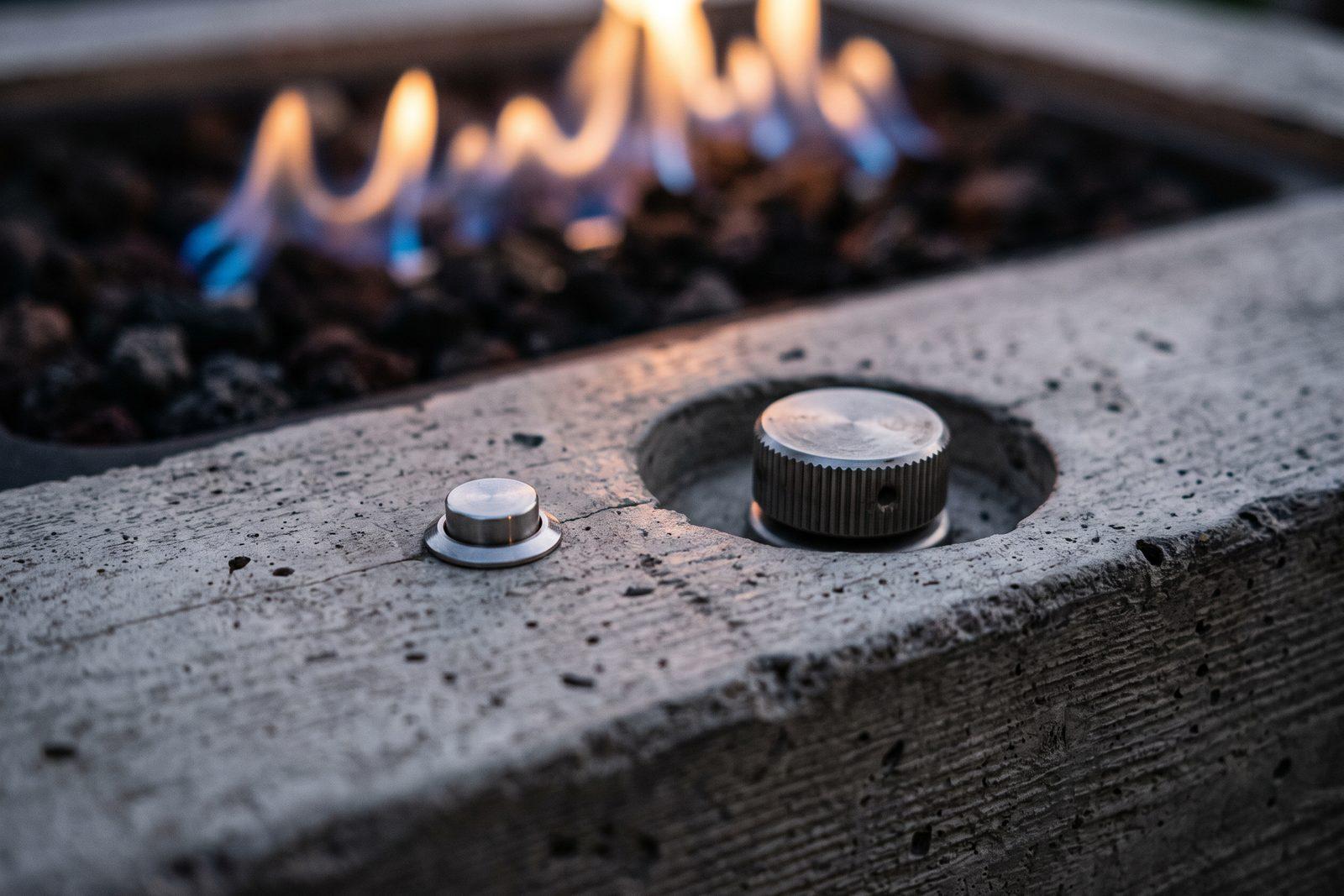 A close view of an electronic ignition control panel set into the edge of a concrete fire table at dusk, lit by warm light from the flame beside it.