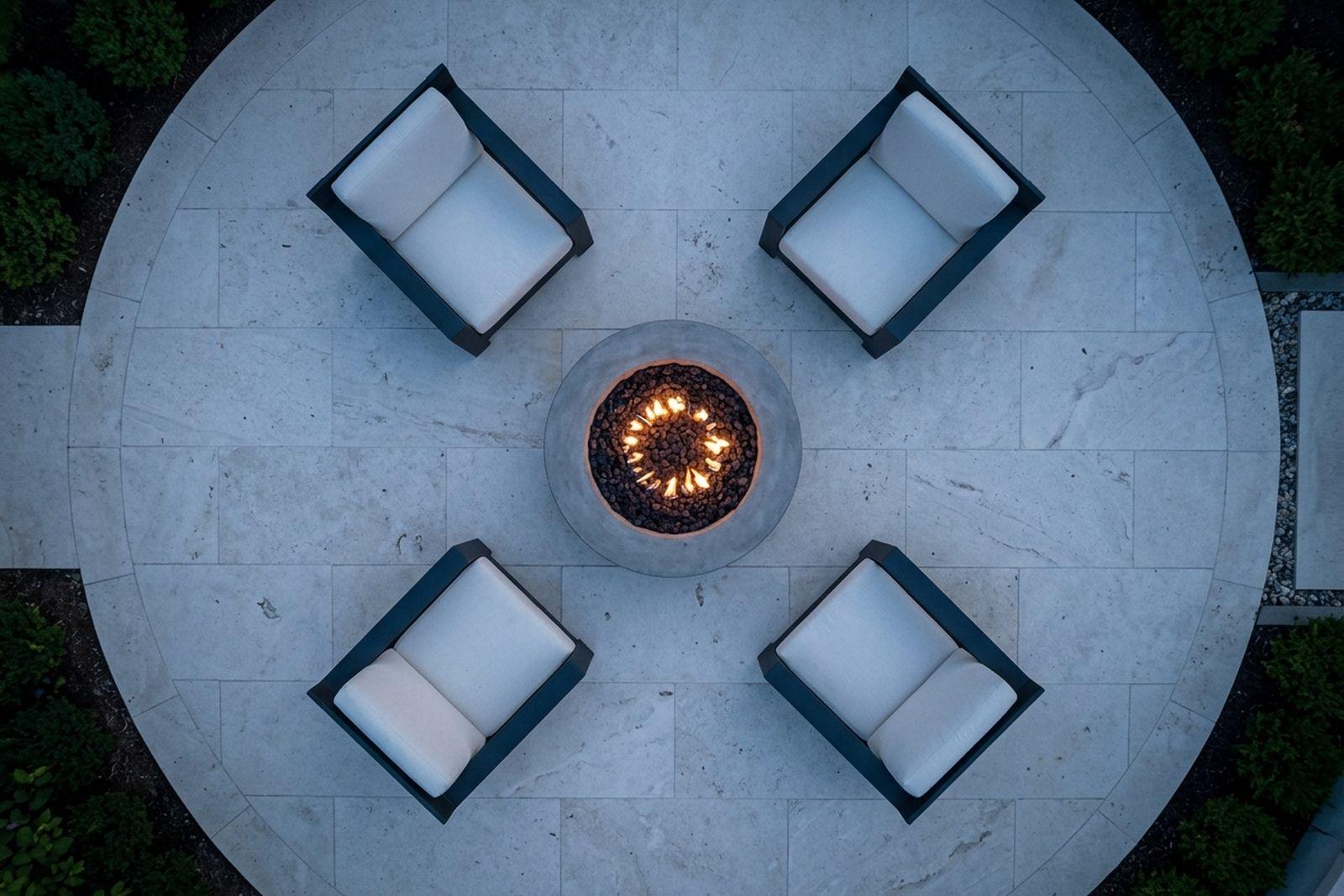Overhead view of a round fire pit ringed by four lounge chairs on a stone patio at dusk.