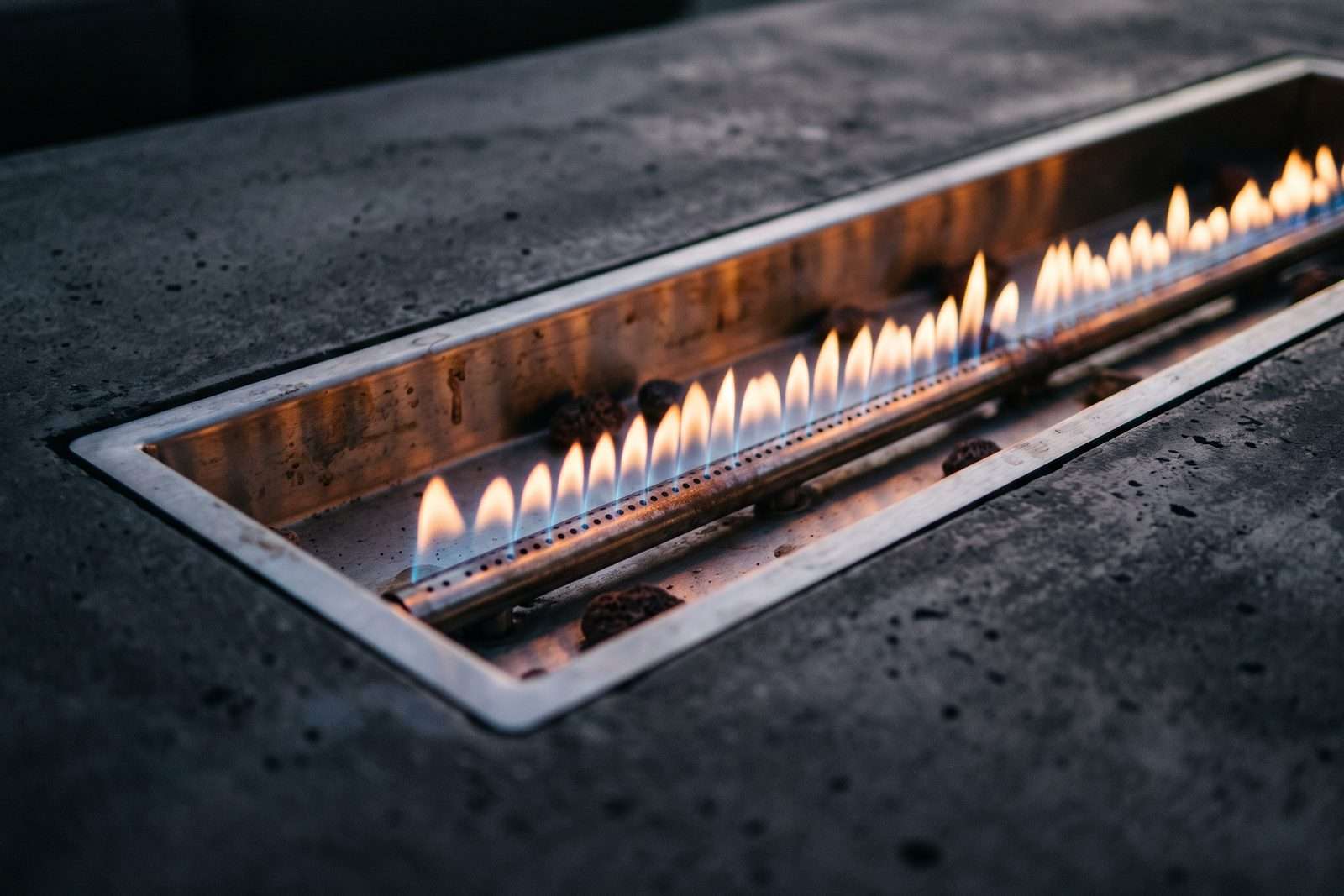 A tight three-quarter view of a linear gas burner flame running along a stainless pan at dusk, the far end of the flame softly out of focus.