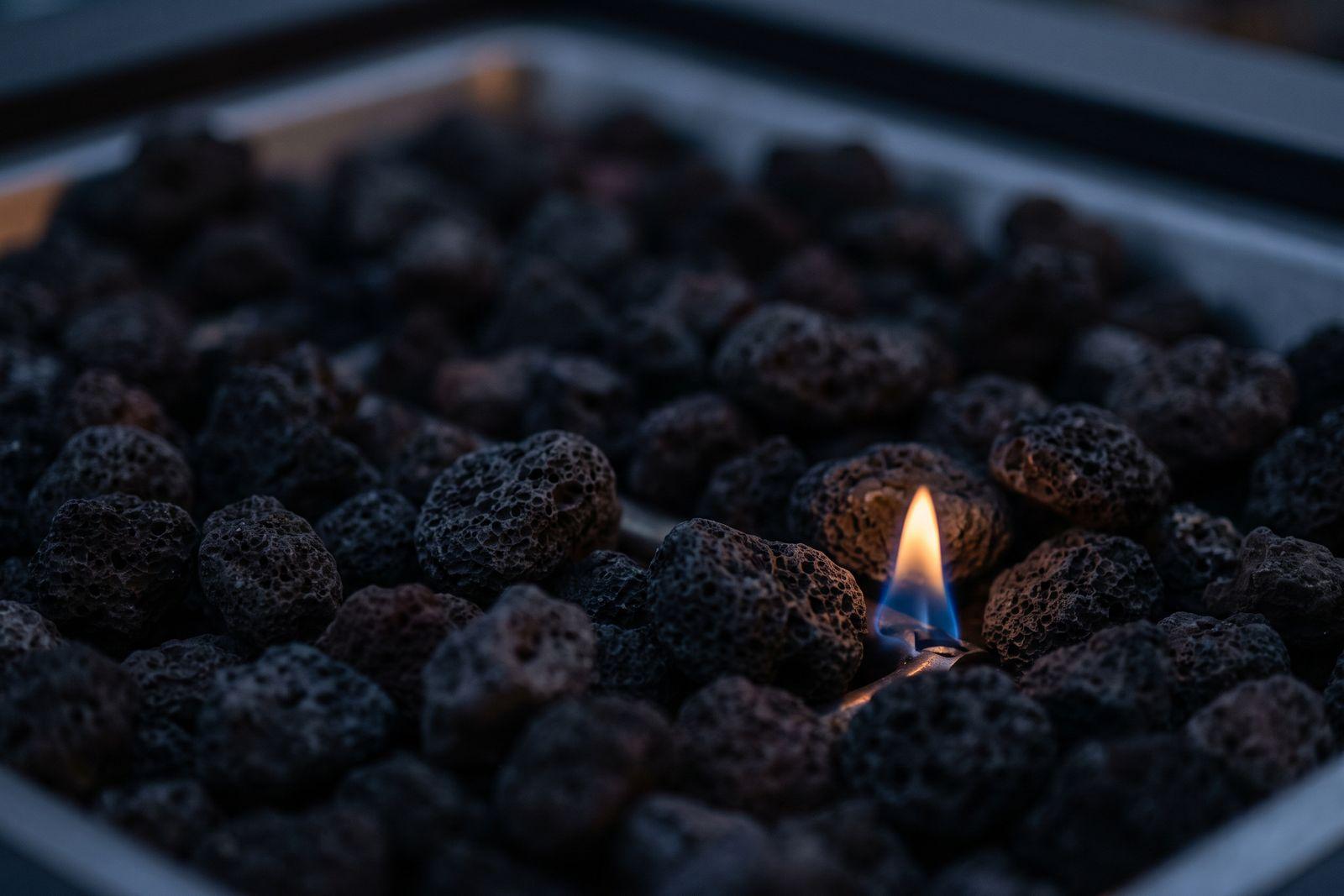 Close detail of a gas fire pit burner with a small flame flickering low over lava stones at dusk.